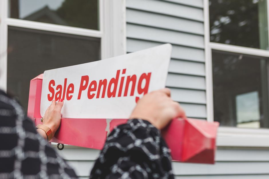 A person is attaching a white and red 'Sale Pending' sign to the front of a modern, grey-clad house. The sign is held firmly with both hands, and the person is wearing a long-sleeved, patterned black-